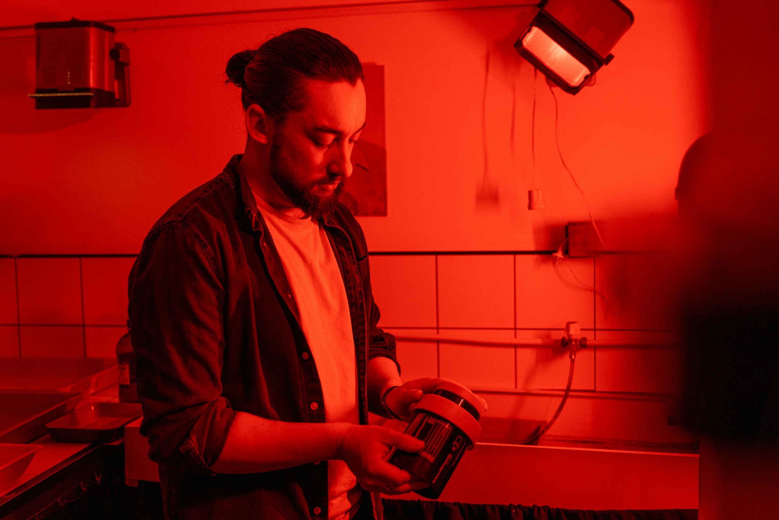 Photographer processing film in a red-lit darkroom, holding developing tank.
